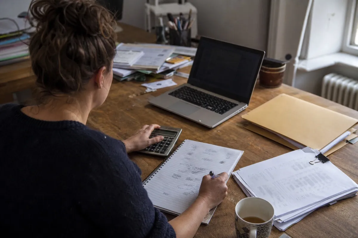 A bookkeeper working out what to charge a client, tapping figures into a desk calculator beside handwritten notes and a stack of paperwork.