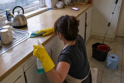 A cleaner in rubber gloves wiping down a kitchen worktop while a phone lights up unanswered nearby