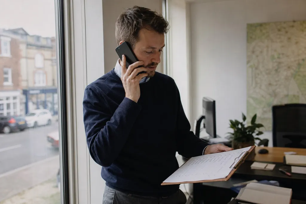 A real estate agent making a follow-up call by the office window with a list of contacts on a clipboard