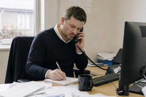 A real estate agent at their desk taking a buyer inquiry on the phone and jotting down notes