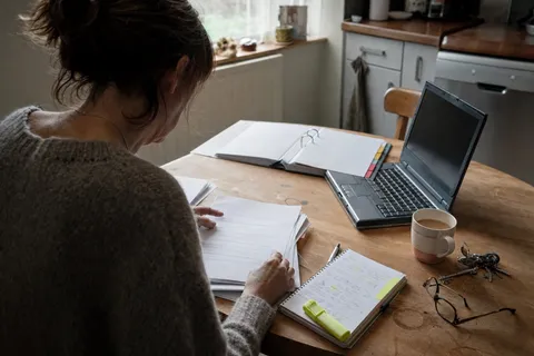A new home care agency owner working through registration forms and policy folders at a kitchen table with a laptop and a mug of tea.