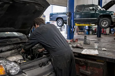 A mechanic leaning under the bonnet of a car in a busy garage while the phone rings unanswered on the cluttered counter behind him