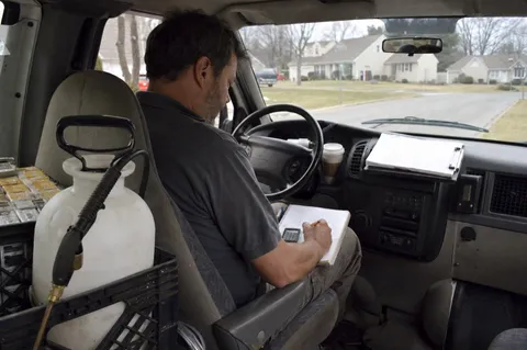 A pest control technician sitting in his work van, writing figures in a notebook with a calculator to work out the price for a job.