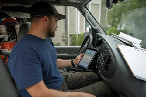 An HVAC technician checking a job-management app on a tablet beside a work truck