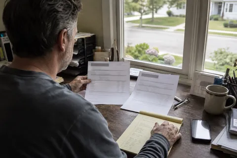 A small business owner at a desk comparing two printed answering service quotes side by side while a cell phone lights up with an incoming call, weighing an AI receptionist against a human answering service.